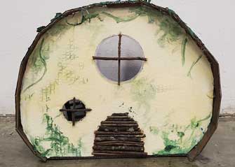 A small cardboard house in the shape of a semi-circle. The walls are painted pale yellow and there's fake greenery and moss added. Theres a round door made with sticks and a circular window above.